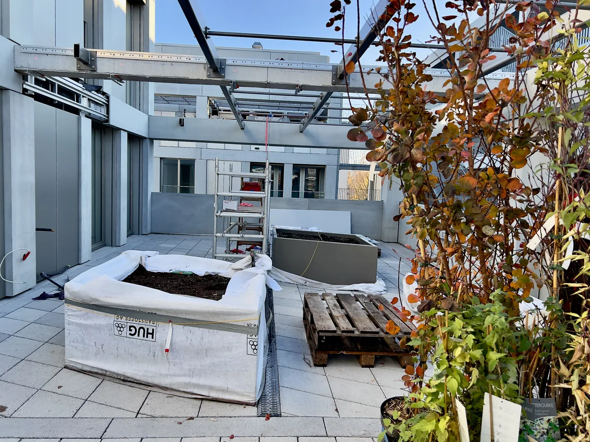 Man sieht eine Terrasse, auf der zwei fest eingebaute Hochbeete stehen. Im Vordergrund sind ein paar Kletterpflanzen zu sehen.